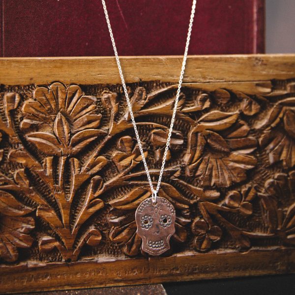 small matt copper stamped sugar skull pendant on a sterling silver chain. Hung on a dark red old fashioned book over a carved wooden panel