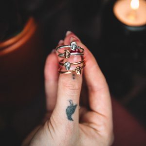 tattoed hand holding 4 mini skull rings in copper and silver with a candle and bowl on a dark background