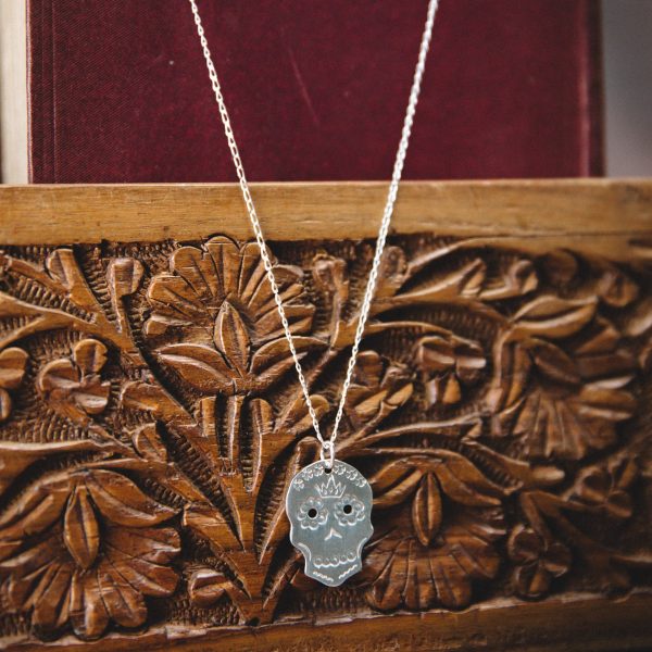 small silver stamped sugar skull pendant on a sterling silver chain. Hung on a dark red old fashioned book over a carved wooden panel
