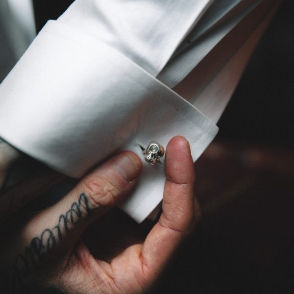 white shirt sleeve being held at the cuff by a young white man with tattoos, small sterling silver skull cufflink in the cuff.