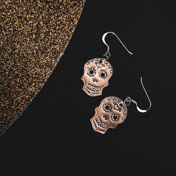 matt copper stamped sugar skulls earrings with earring wires laid on a black leather mat placed underneath with a copper mat beside them