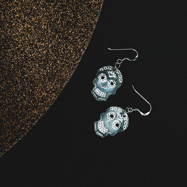 silver stamped sugar skulls earrings with earring wires laid on a black leather mat placed underneath with a copper mat beside them