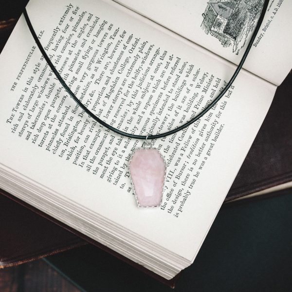 rose quartz coffin necklace made from sterling silver and a black thong necklace laid on a open old fashioned book with other books beneath