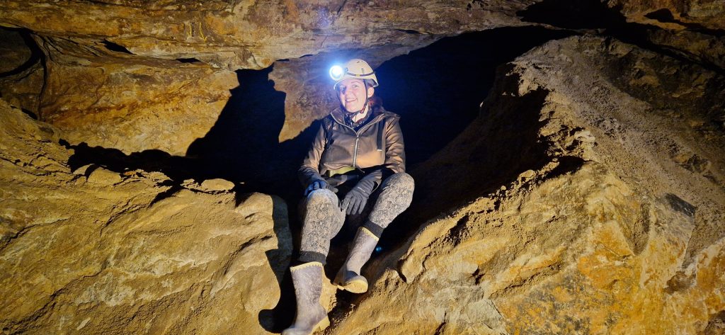 white woman in a bronze jacket and brown leggings and purple wellies sitting on a sandstone ledge in a mine with a hard hat and torch smiling.