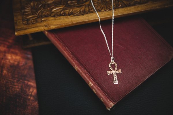 patinated copper ankh necklace with a sterling silver chain laid on top of a dark red old fashioned book with a wooden background