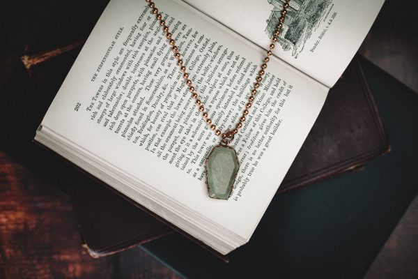 pale green jade coffin necklace made from copper with a copper ball chain necklace laid on a open old fashioned book with other books beneath
