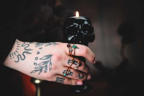 tattooed hand holding a black skull candle holder with 10 different gemstone copper rings on 3 different fingers on a dark background