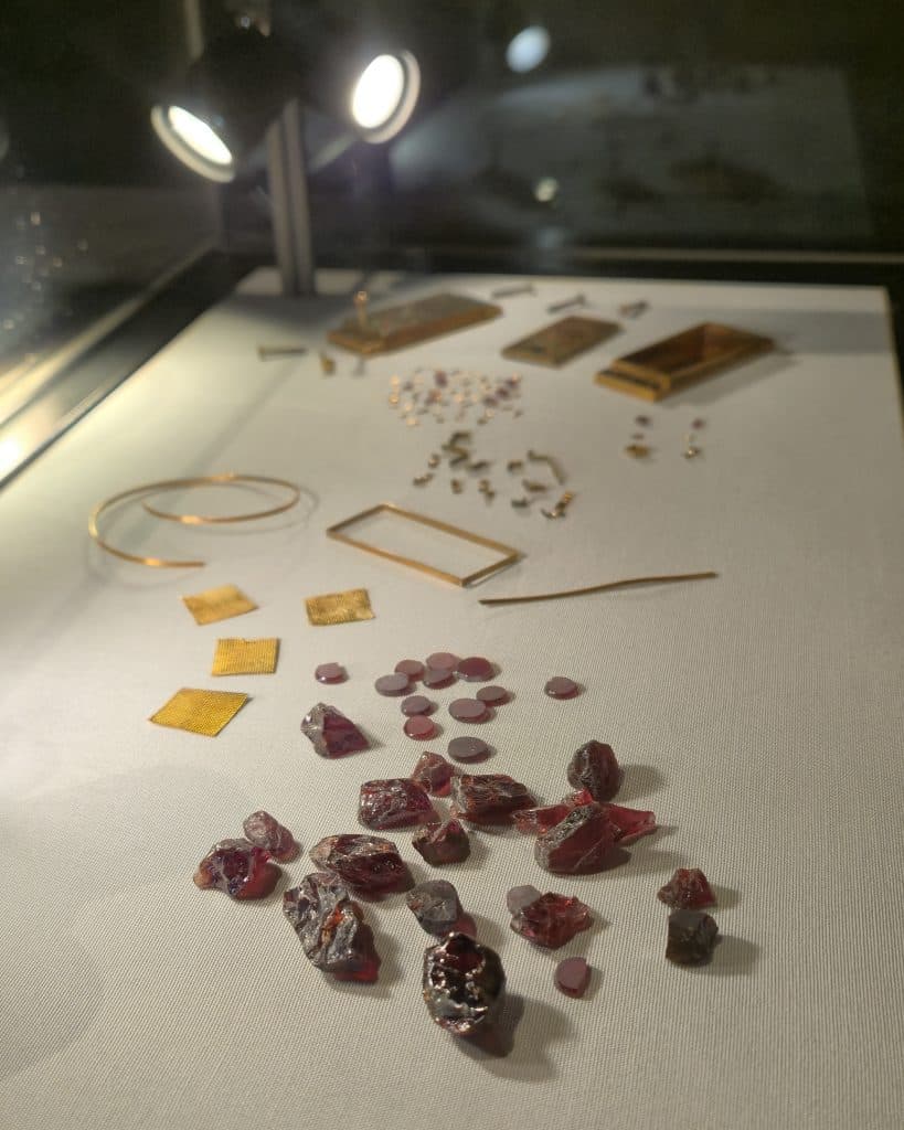 scattered rough red garnet stones in a display case in front of pieces of gold