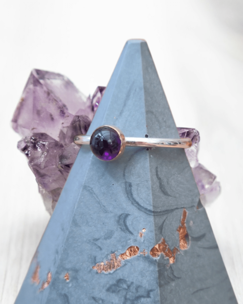 amethyst cabochon stone ring set in rose gold bezel with sterling silver band on a concrete style ring cone in front of an amethyst crystal