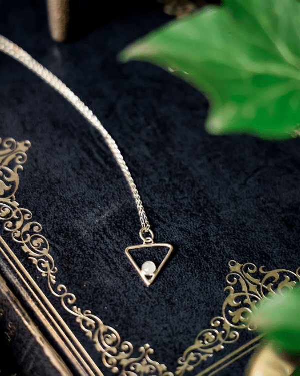 upside down triangular shaped silver pendant with pale blue stone set in bottom point. On a silver chain laid on a navy blue antique hardback book with ivy leaves