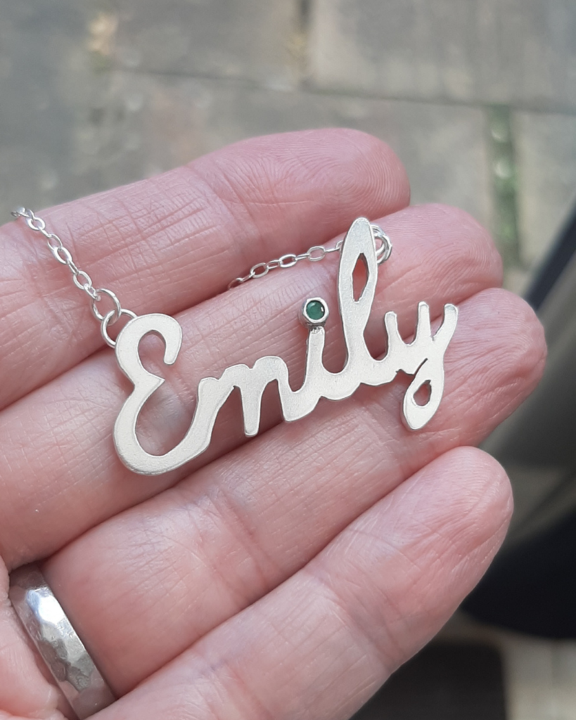 Silver necklace in the name of Emily with emerald set in "i" in a woman's hand