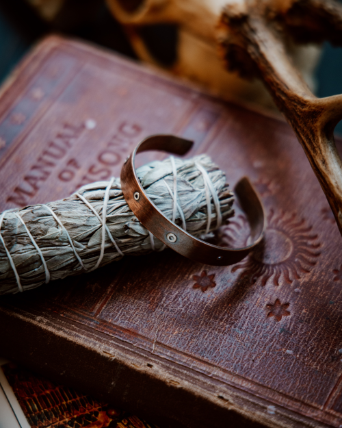 copper cuff with silver rivets sitting on a sage stick on old books