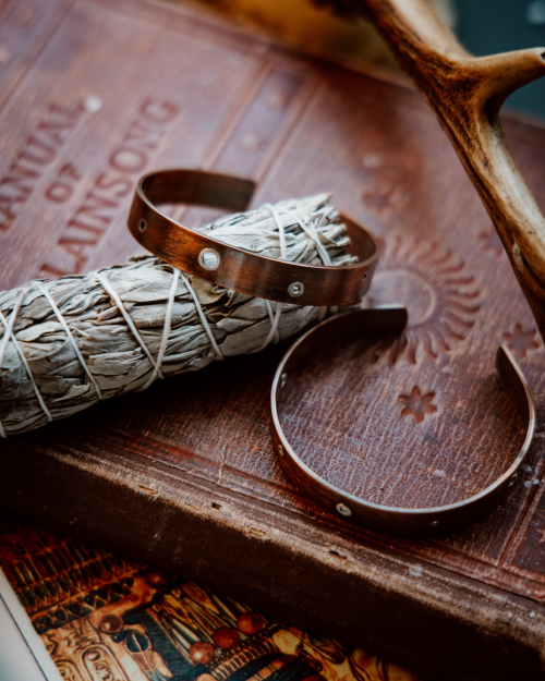 2 copper cuffs with silver rivets sitting on a sage stick on old books