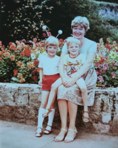 a woman and two children sat on a stone wall with flowers behind them smiling at the camera