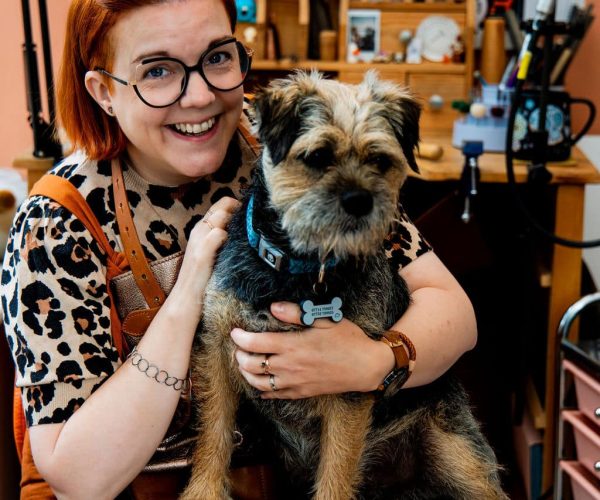 smiling red haired lady sat in front of a jewellers bench with a border terrier on her lap