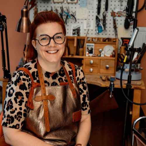 smiling red haired lady in a copper leather apron sat in front of a jewellery bench