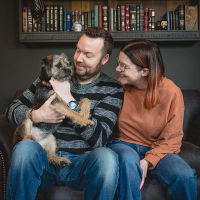 sam, ed and boba the border terrier sat together on their grey sofa in front of their book case in their lounge
