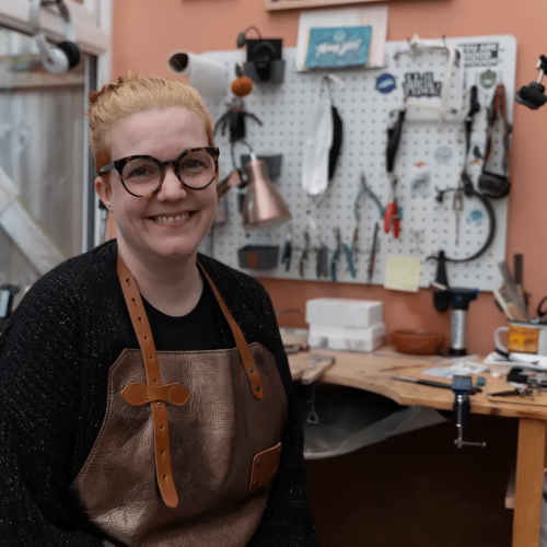 woman with glasses and hair tied up smiling at the camera sat in front of a jewellery bench with tools.